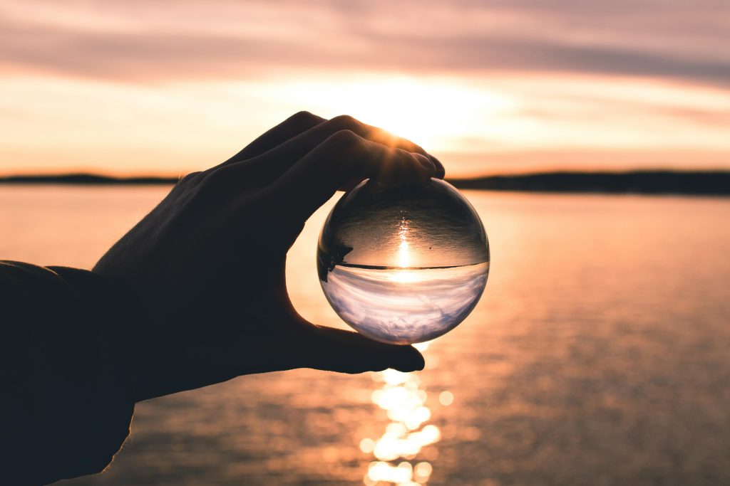 A hand holds a crystal ball reflecting a serene sunset over calm ocean waters.